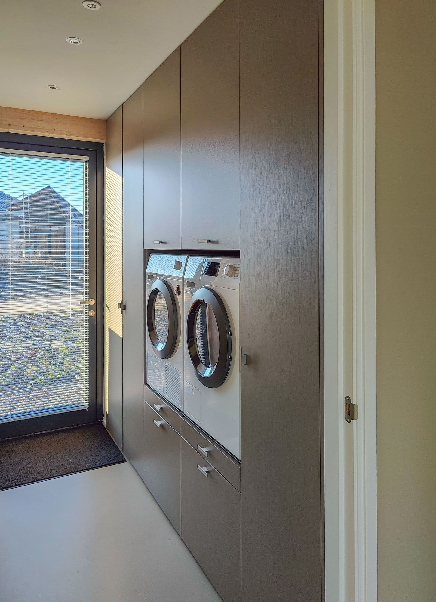  Voorzijde van een moderne wasmachinekast met ingebouwde wasmachine en droger, en opbergruimte erboven, geplaatst in een bijkeuken in Almere.