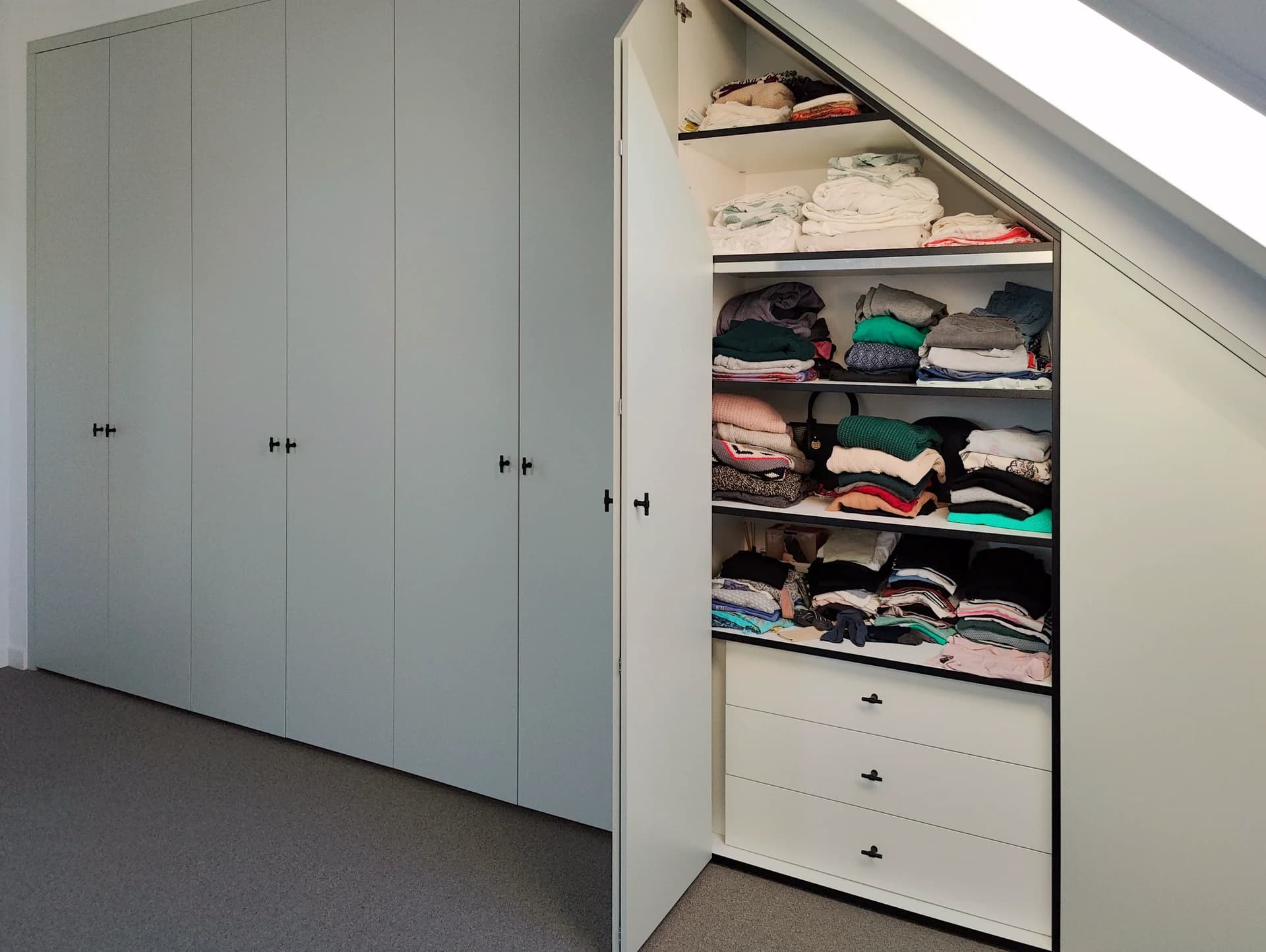 Ingebouwde kledingkast onder schuin dak met geopende deuren, toont overzicht van planken en laden.