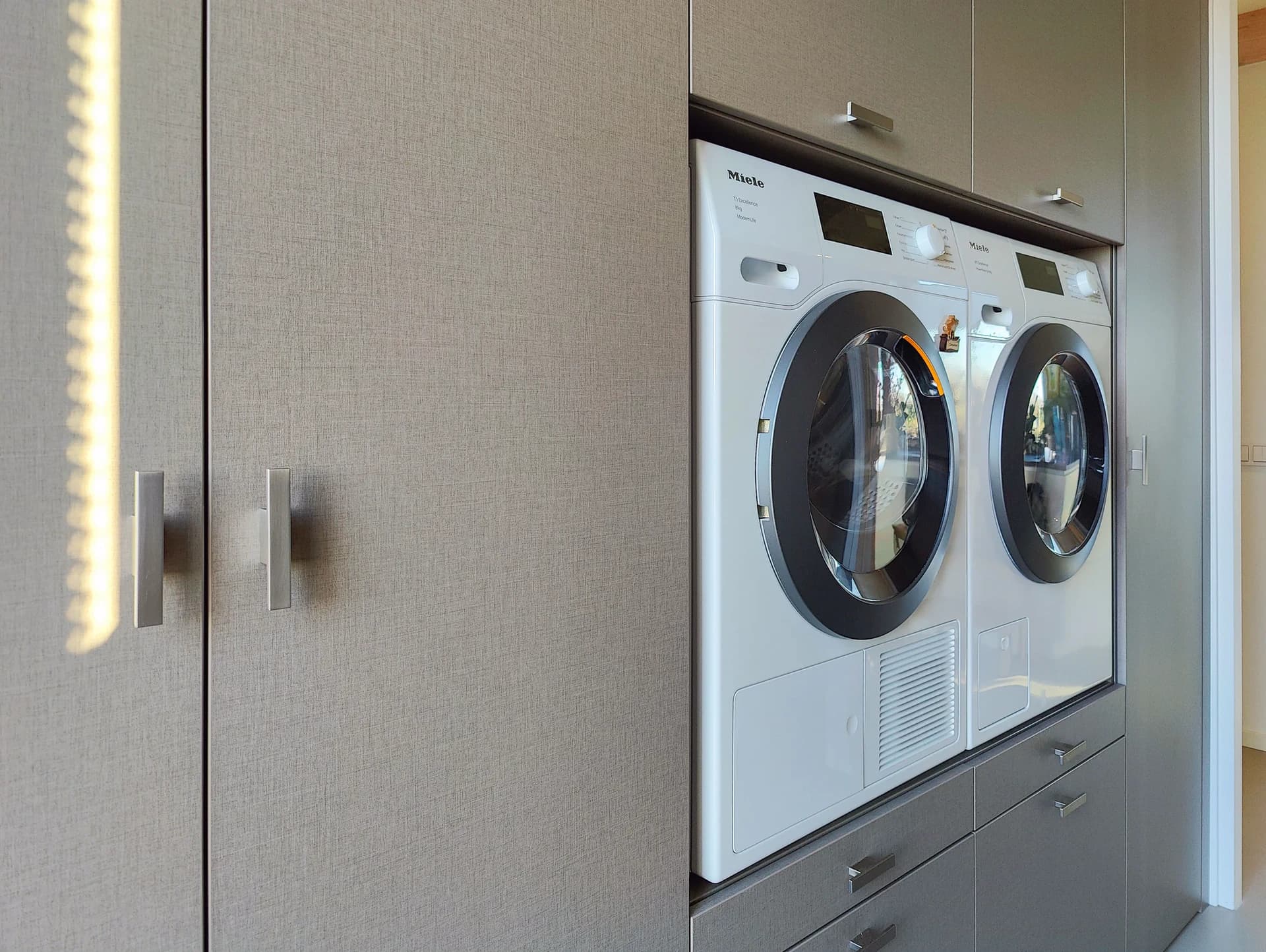 Voorzijde van een moderne wasmachinekast met ingebouwde wasmachine en droger, en opbergruimte erboven, geplaatst in een bijkeuken in Almere.